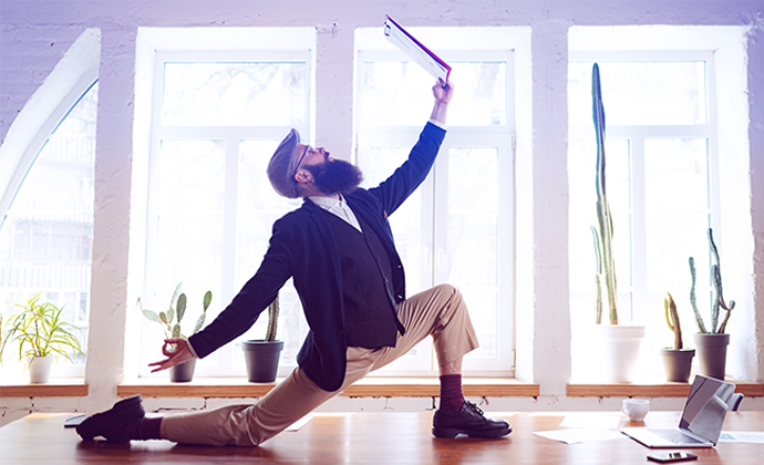 Business professional striking a dynamic pose on a desk with a laptop and documents, symbolising balance and a healthy business mindset.
