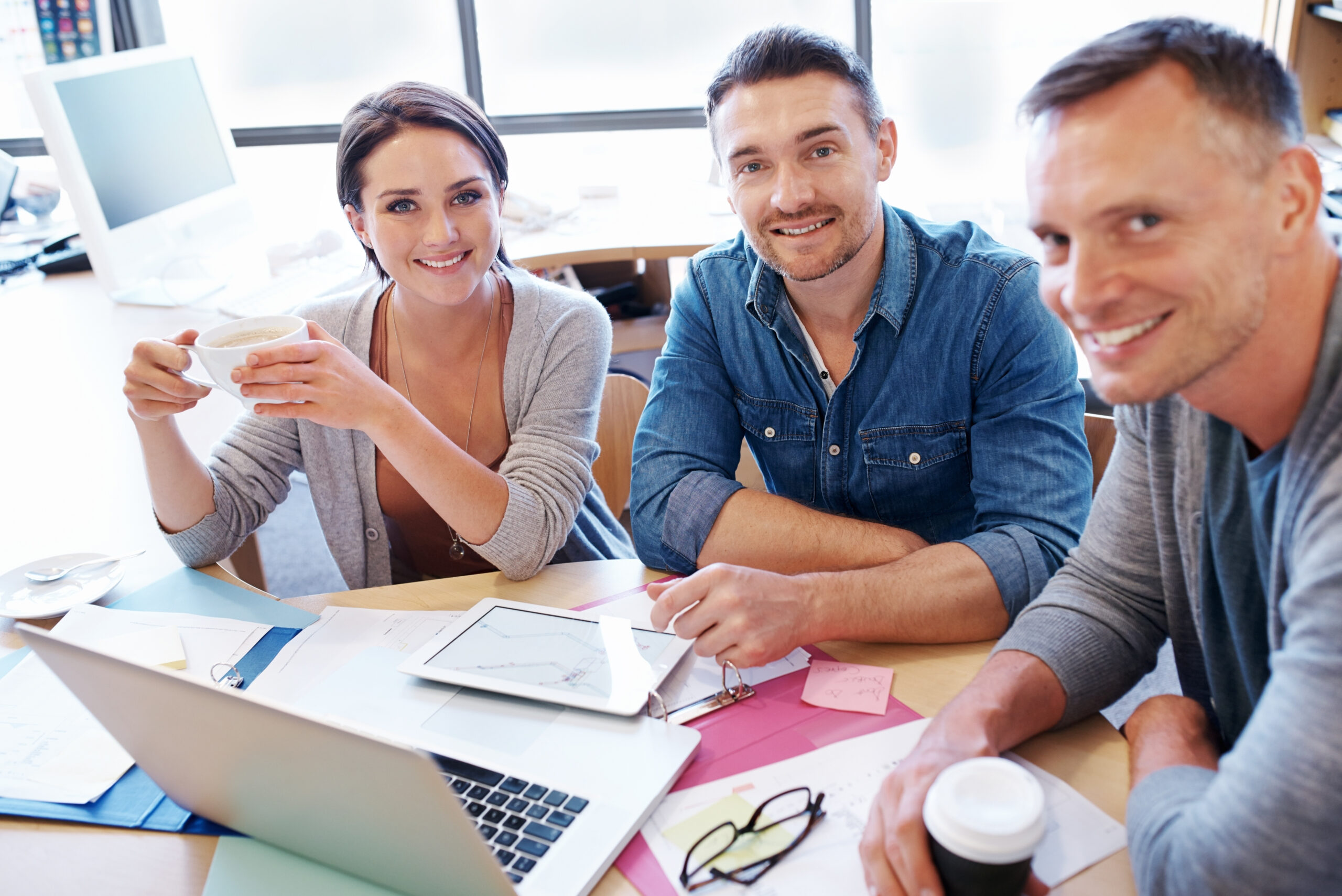 Three professionals sit at a desk smile at a laptop